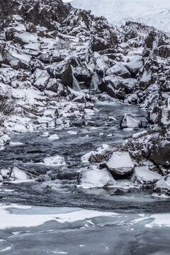 View of a fast-flowing river cuts through the snow-dusted, rocky terrain, creating a stark contrast between the dark stone and icy water, Iceland.