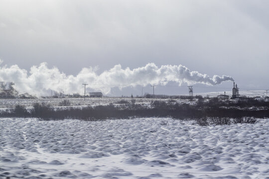View of the stark winter landscape with snow-covered ground, a distant industrial complex emitting plumes of smoke under a cloudy sky, Iceland.