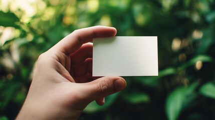 A close-up of a human hand holding a blank white rectangular card against a softly blurred green foliage background.  Perfect for branding mockups, presentation templates 