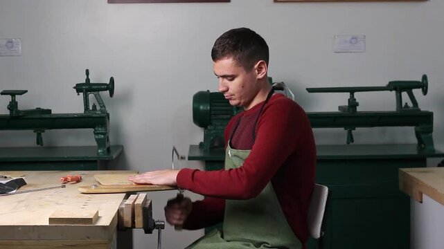 Young craftsman in apron filing or rasping wood piece at workbench in workshop with lathes. Focused woodworking process. A man cuts plywood with a jigsaw in a workshop