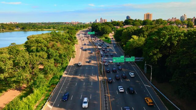 Aerial view of Interstate 678 with multi-lane traffic in New York City boroughs, Queens. Road among green trees of a park on the shore of a lake on a sunny day.