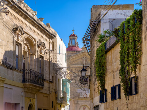 Attard, Malta &ndash; narrow old town alleyway with a view of the parish church and beautiful old town facades with hanging plants and warm sunlight