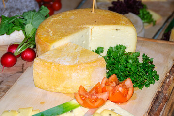 Rustic cheese wheel with a cut wedge on a wooden board, garnished with fresh parsley, tomatoes and vegetables, ideal for gourmet food, dairy and market themes.