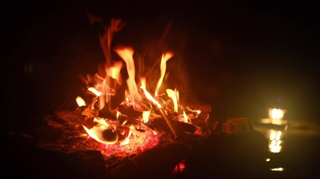 Relaxing night campfire near river with light of lantern. Beautiful bonfire, burning old wood in autumn or summer time. Active lifestyle, traveling, hiking. Camping vibes and outdoor lifestyle mood