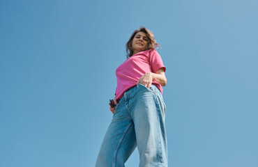 Beautiful carefree young woman with curly hair in pink t-shirt smiling and looking down at camera on blue sky background low angle shot.