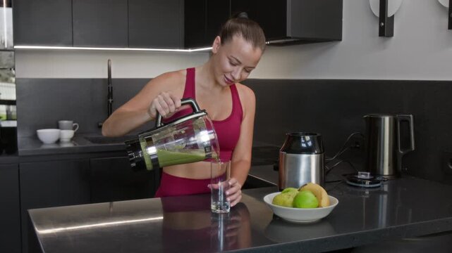 Cheerful young woman removing jar off blender and pouring green smoothie into glass after preparing morning drink on kitchen countertop
