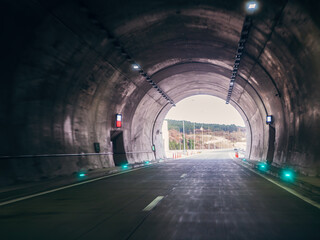 Concrete road tunnel with LED lights leading to bright highway exit surrounded by forested landscape.