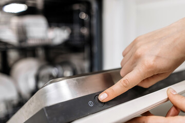 Close-up of a hand pressing the start button on a modern dishwasher, showcasing sleek design and functionality in a contemporary kitchen environment with clean dishes