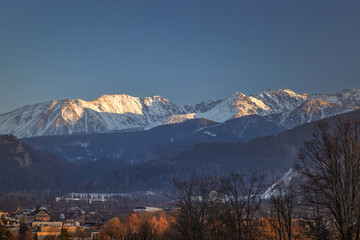 Snow-capped mountains rise majestically over a city nestled among rolling hills and forests under a clear sky. Zakopane town at the foot of the Tatra Mountains in Poland, Europe.
