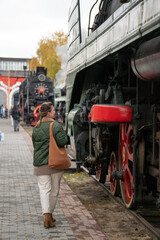 Woman in a green jacket stands beside a vintage steam locomotive with red wheels on an outdoor railway platform, industrial details and autumn mood