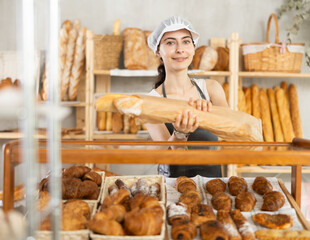 Young Armenian female employee in black apron sells fresh baguettes made of white flour packed...