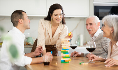 Obraz premium Joyful senior parents and young couple sitting around table playing brick game and drinking wine in goblets at home