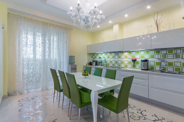 A kitchen with a large white table and green chairs has bright lights and a light ambiance. There are colorful tiles on the wall and flowers on the counter