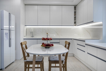 In a bright kitchen, there are white cabinets and a round table with a bowl of apples. Two wooden chairs are placed around the table. A refrigerator is nearby