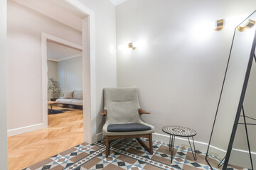 A hallway features a simple chair, a round table, and a mirror mounted on the wall. Light fixtures provide illumination. The space shows sleek design. A living room is visible in the background