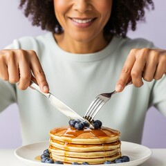 Smiling woman cuts into a stack of pancakes with blueberries and syrup on a white plate at the table.