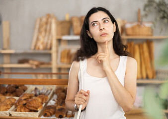 Young female visitor stopped near display case with pastry, examines, view and chooses sweet snack croissants, makes order, bagel, crescent rolls, pastry in bakery