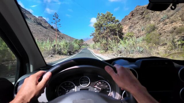 First person view of a man driving a minivan on a curvy road through a scenic volcanic canyon in Gran Canaria, surrounded by lush vegetation under a clear blue sky on a sunny summer day