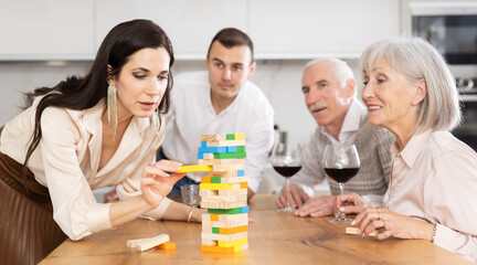 Group of mature spouses and middle-aged male visitor with girlfriend is playing board game. Senior married couple build tower with wooden blocks bricks with younger spouses