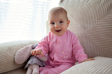 Happy infant baby girl playing on couch at home wearing pink bodysuit. Cute child smiling.