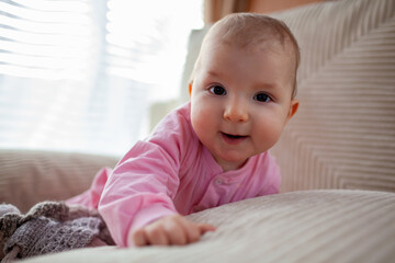 Happy infant baby girl playing on couch at home wearing pink bodysuit. Cute child smiling.