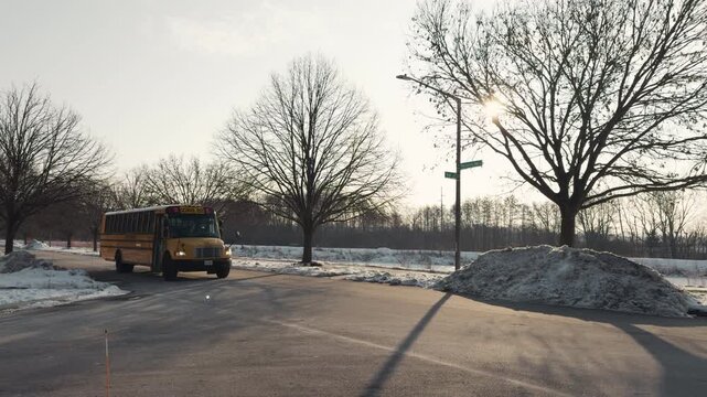 Yellow school bus slowly departing a stop and pulling away along a suburban street. Snowbanks line the roadway as the bus slowly departing winter conditions. POV View