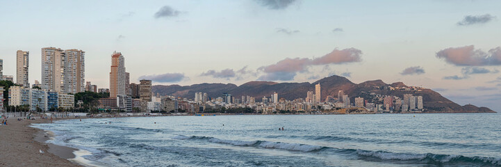 Sunset view of Benidorm from Poniente beach, unrecognizable people swimming in the sea and relaxing on the sand, city buildings along the coast, and a mountain with colorful clouds in the background.