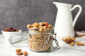 Tasty granola with dried fruits in glass jar on white marble table, closeup