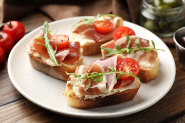 Delicious bruschettas with cream cheese, prosciutto, tomatoes and arugula on wooden table, closeup