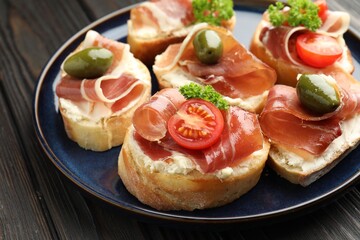 Delicious bruschettas with cream cheese, prosciutto, green olives, tomatoes and parsley on dark wooden table, closeup
