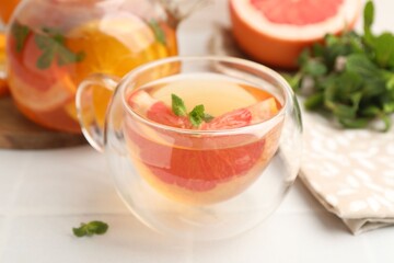 Aromatic fruit tea in glass cup, teapot, grapefruit and mint on white table, closeup