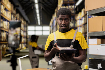 Black male employee vlogging with tripod for a commercial in industrial warehouse, presenting the large scale import export operations on a video for advertisement. Creating promotional content.