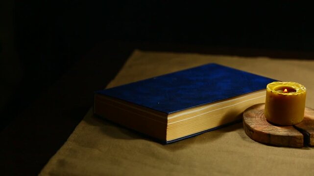 Burning wax vintage candle with blue book on the  table on dark black background with copy space. Ancient old education creative idea concept in candle light