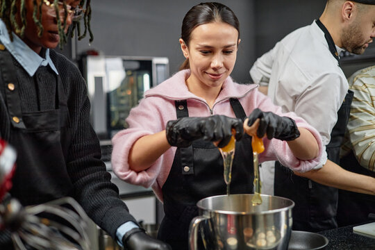 Young adult Caucasian woman cracking eggs into mixing bowl while participating in cooking class with multiethnic group of adults, chef instructing in background, kitchen setting