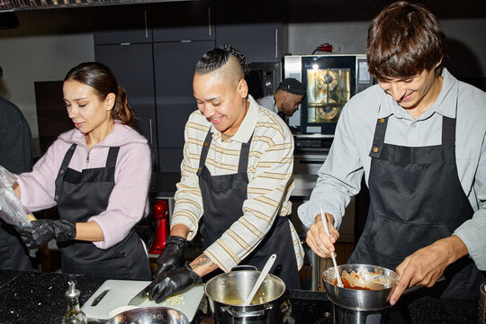 Multiethnic group of young adults cooking together in kitchen, Caucasian man stirring mixture in bowl, biracial woman chopping vegetables, young woman preparing ingredients, all wearing aprons