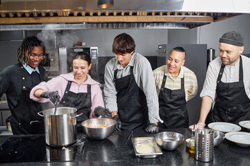 Group of young multiethnic men and women cooking together in professional kitchen, smiling and preparing food in large pot during culinary class