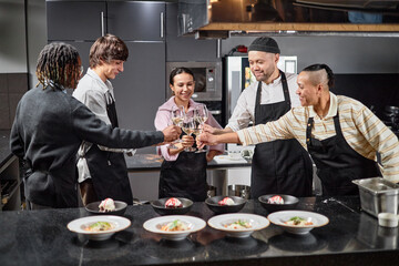 Group of multiethnic young adults wearing aprons standing in kitchen clinking glasses together celebrating teamwork with plated dishes on counter