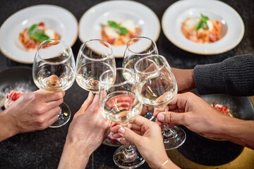 Diverse group of young hands clinking wine glasses during celebratory meal, visible plates of food in background, multiethnic gathering sharing toast