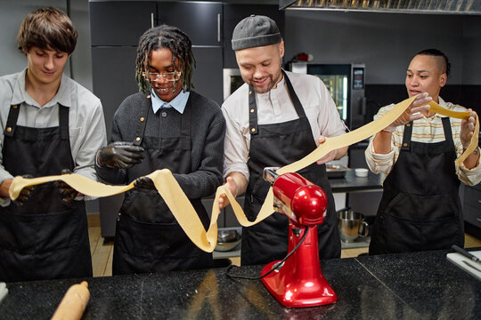 Group of young adult woman and men, including Black and Caucasian individuals, making fresh pasta together in kitchen, holding long sheet of dough coming from electric pasta machine