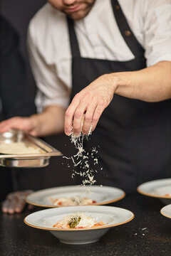 Caucasian young adult man sprinkling grated cheese over plated pasta dishes in kitchen, wearing apron and chef uniform, focusing on food preparation with visible hand motion