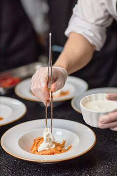 Man preparing pasta dish, using tongs to place creamy sauce on noodles, wearing gloves, working in professional kitchen with plates in background