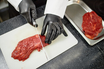 Chef wearing black gloves slicing raw beef steak on cutting board with stainless steel knife, preparing meat for cooking, second piece of beef resting on metal tray nearby