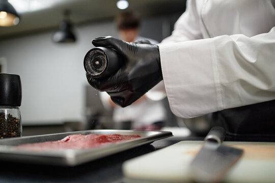 Chef wearing black gloves seasoning raw meat with pepper grinder in professional kitchen, focusing on preparing ingredients for cooking, coworker visible in background