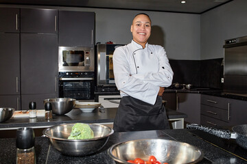 Portrait of young biracial chef standing with arms crossed in professional kitchen, smiling confidently, stainless steel bowls with vegetables and kitchen appliances visible
