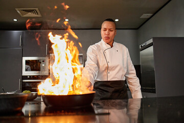 Biracial female chef cooking with open flame in professional kitchen, standing behind stove and concentrating on preparing food in skillet, modern appliances visible