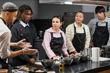 Diverse group of young adults listening to chef instructor demonstrating cooking techniques in kitchen classroom, all wearing aprons and standing by counter