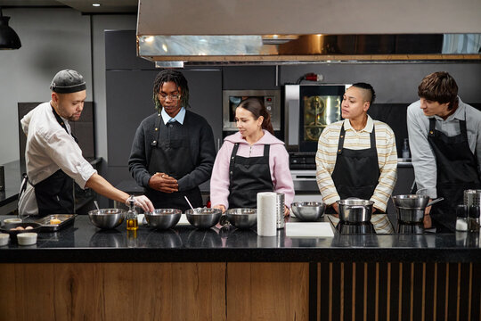 Chef instructing group of diverse people in kitchen, demonstrating cooking technique while group attentively observing and wearing aprons at counter