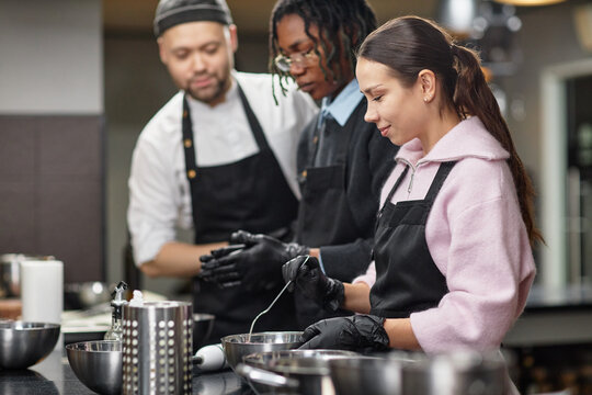 Young Caucasian woman, Black man, and Caucasian man preparing food together in kitchen, wearing aprons and gloves, collaborating while mixing ingredients in metal bowls
