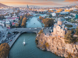 Metekhi Church on a cliff overlooking the Kura River and Tbilisi city skyline, featuring bridges and a cityscape at sunset © EdNurg