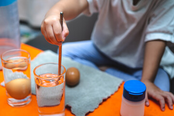 Close-up of a child's hand using a metal chopstick to stir salt into a glass of water on an orange table. Concept of a home science experiment, dissolving solids, and early STEM education.